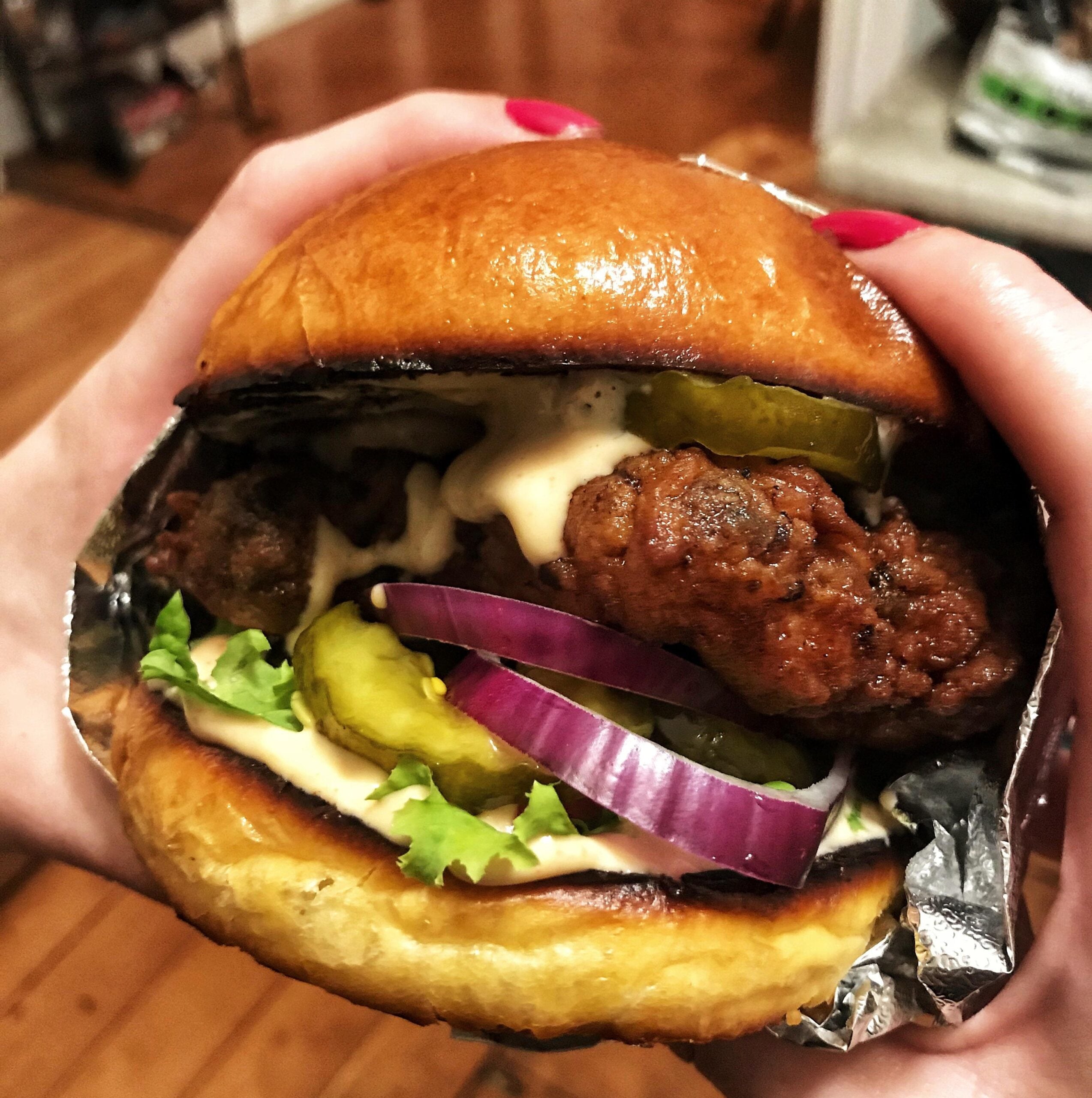 A hand holds a fried chicken sandwich with lettuce, pickles, red onion, and sauce on a toasted bun, partially wrapped in foil.