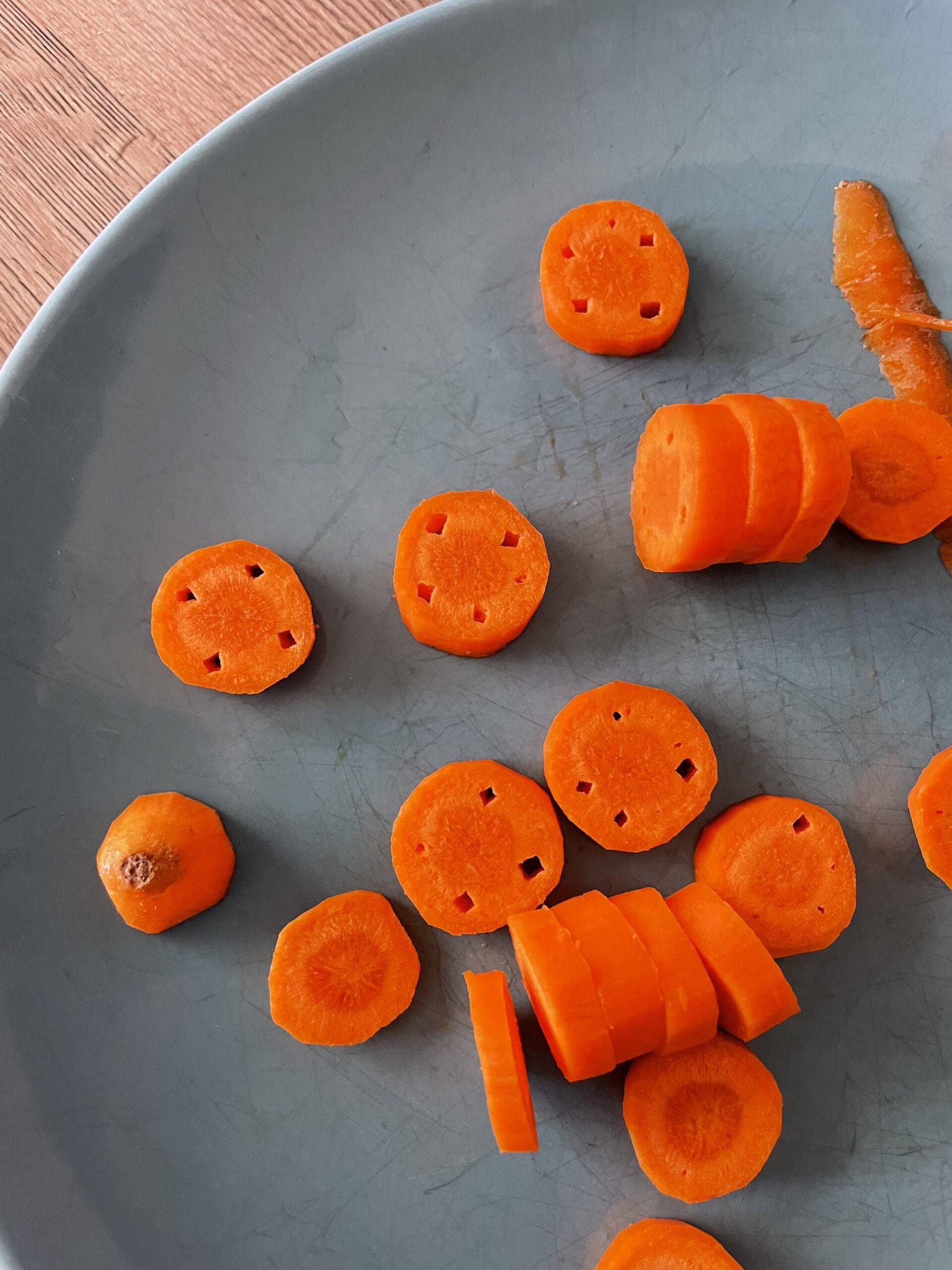 Sliced carrots on a gray plate. Some slices have small square holes arranged in a pattern, while others are solid. A small carrot end is also visible.