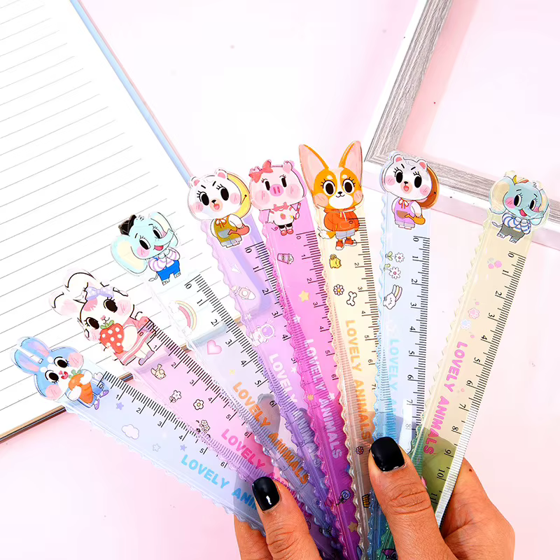 A hand holds seven colorful plastic rulers with cartoon animal characters on top, fanned out. There’s a lined notebook and a silver frame in the background.