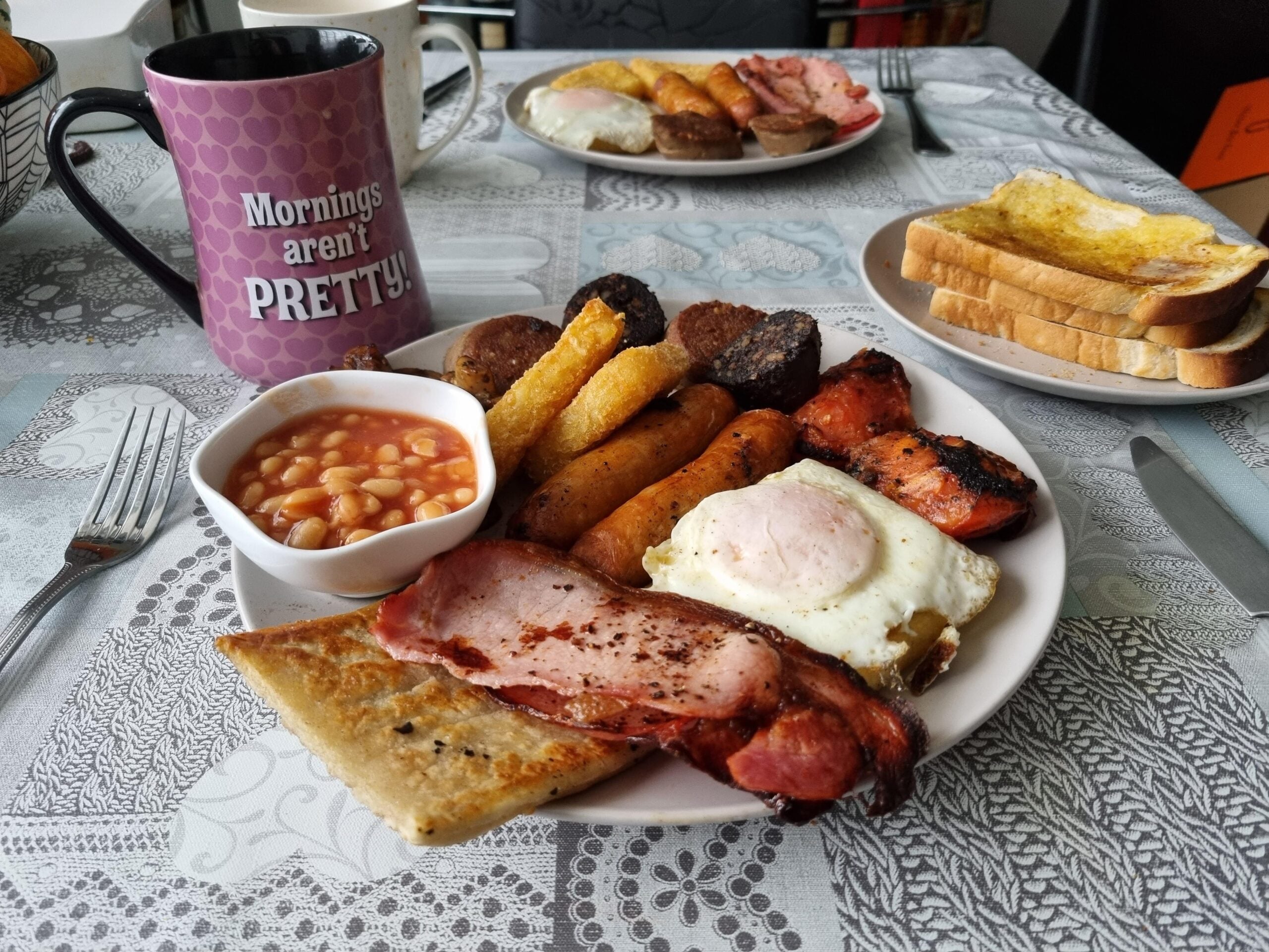 A plate with fried egg, bacon, sausage, black pudding, beans, hash brown, and toast. A mug reading “Mornings aren’t PRETTY!” and two more plates with food and toast are on the table.