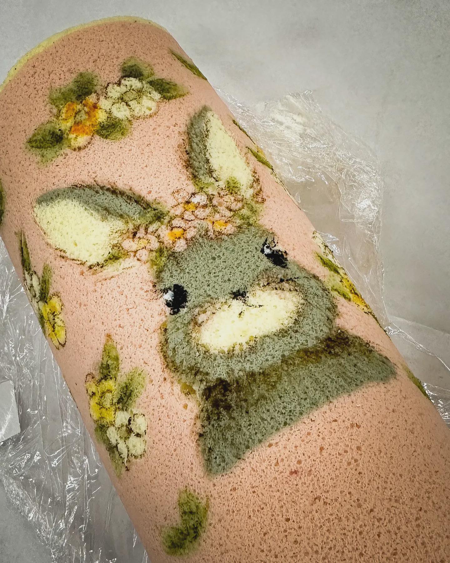 A rolled cake with a pink background features a cute bunny face and yellow flowers, all created using colored cake batter, on a sheet of clear plastic wrap.
