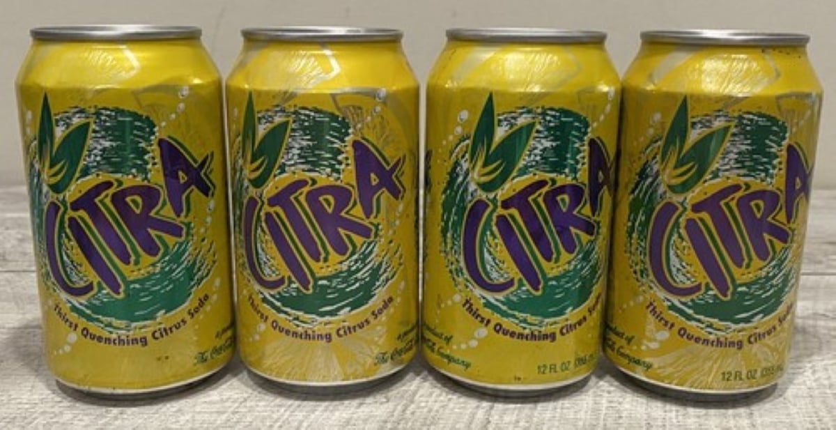Four yellow cans of Citra Soda Pops with green and purple branding are arranged side by side on a light wooden surface.