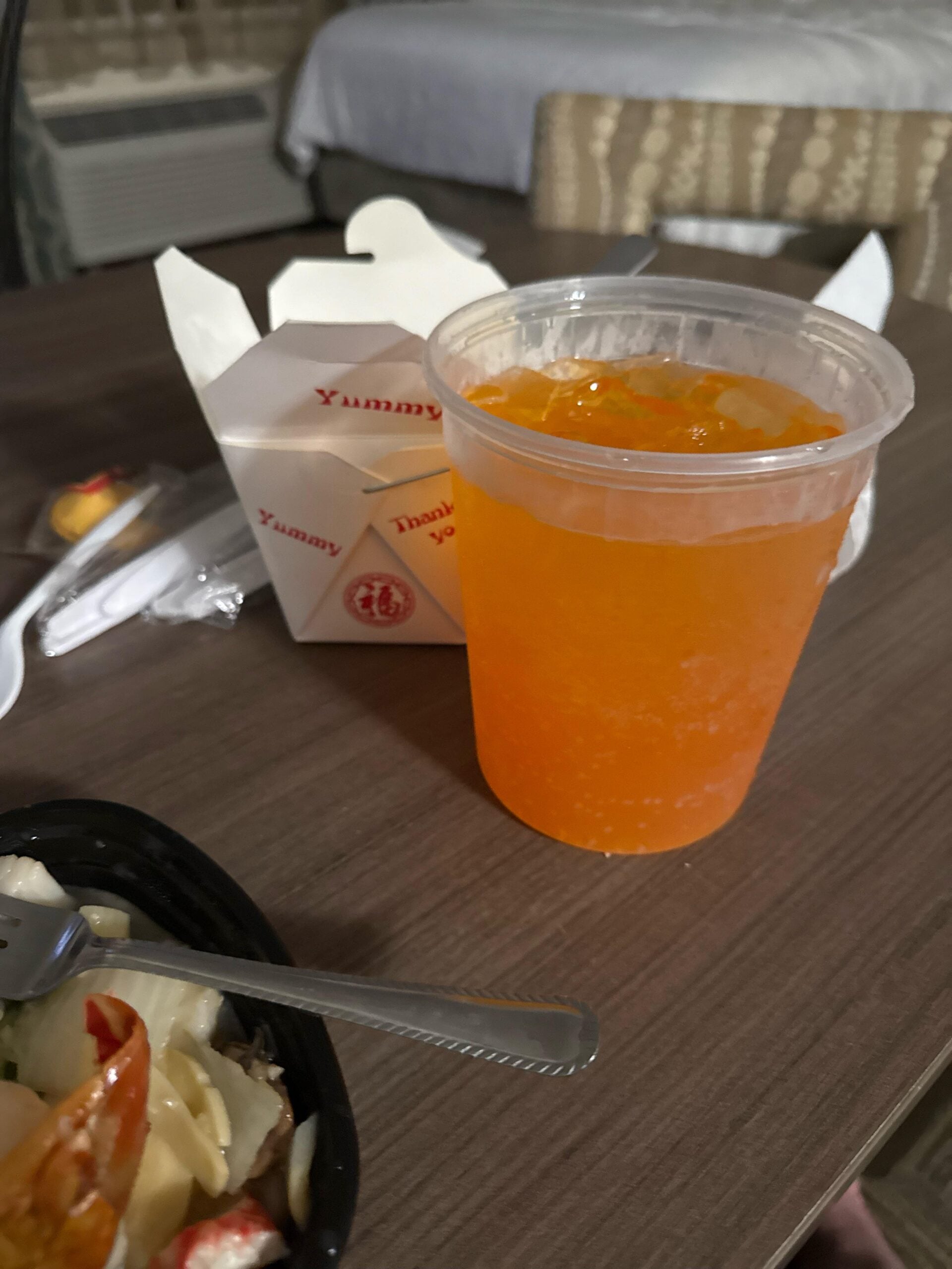 A plastic cup of orange soda sits on a table beside a white takeout box and a partially visible meal in a black container. Disposable utensils and a fortune cookie are also on the table.