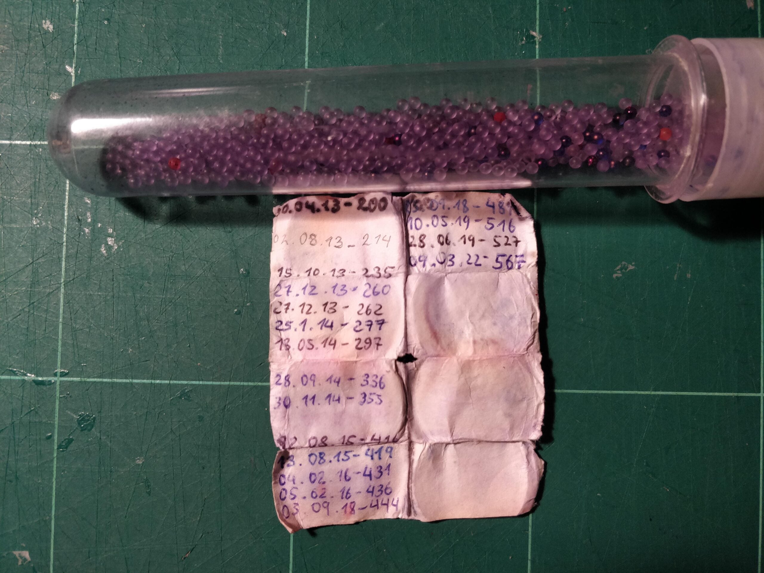 A clear tube filled with purple beads rests on a green cutting mat above folded, handwritten notes listing dates and numbers in blue ink.