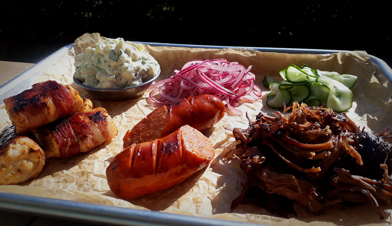 A tray with grilled bacon-wrapped items, sausages, pulled meat, sliced pickles, red onions, cucumber, and a small dish of creamy salad, all on parchment paper.