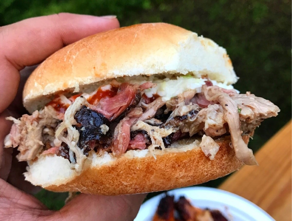 A hand holds a pulled pork sandwich with barbecue sauce on a soft bun. The meat appears juicy and tender, with crispy bits mixed in. A blurry plate and green grass are visible in the background.