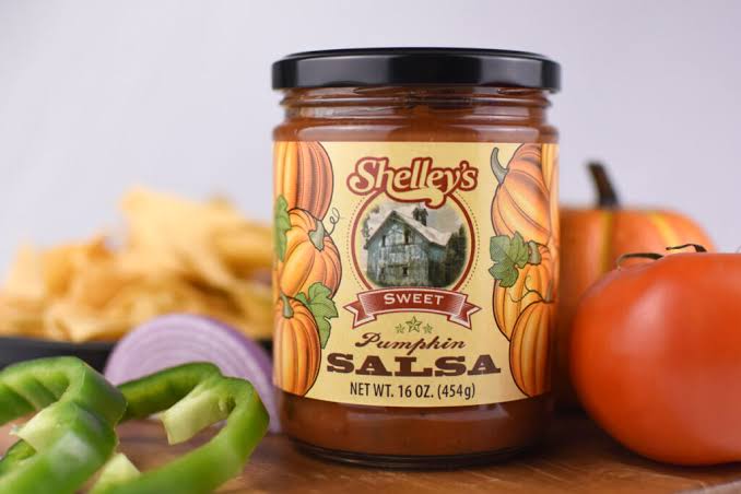 A jar of Shelley’s Sweet Pumpkin Salsa is surrounded by sliced green pepper, a tomato, part of a pumpkin, a red onion, and tortilla chips. The salsa label features pumpkins and an old farmhouse.