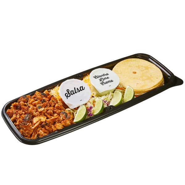 A black tray holding tortillas, seasoned chicken, shredded cheese, cabbage, lime wedges, and two sauce containers labeled “Salsa” and “Cilantro Lime Crema.”