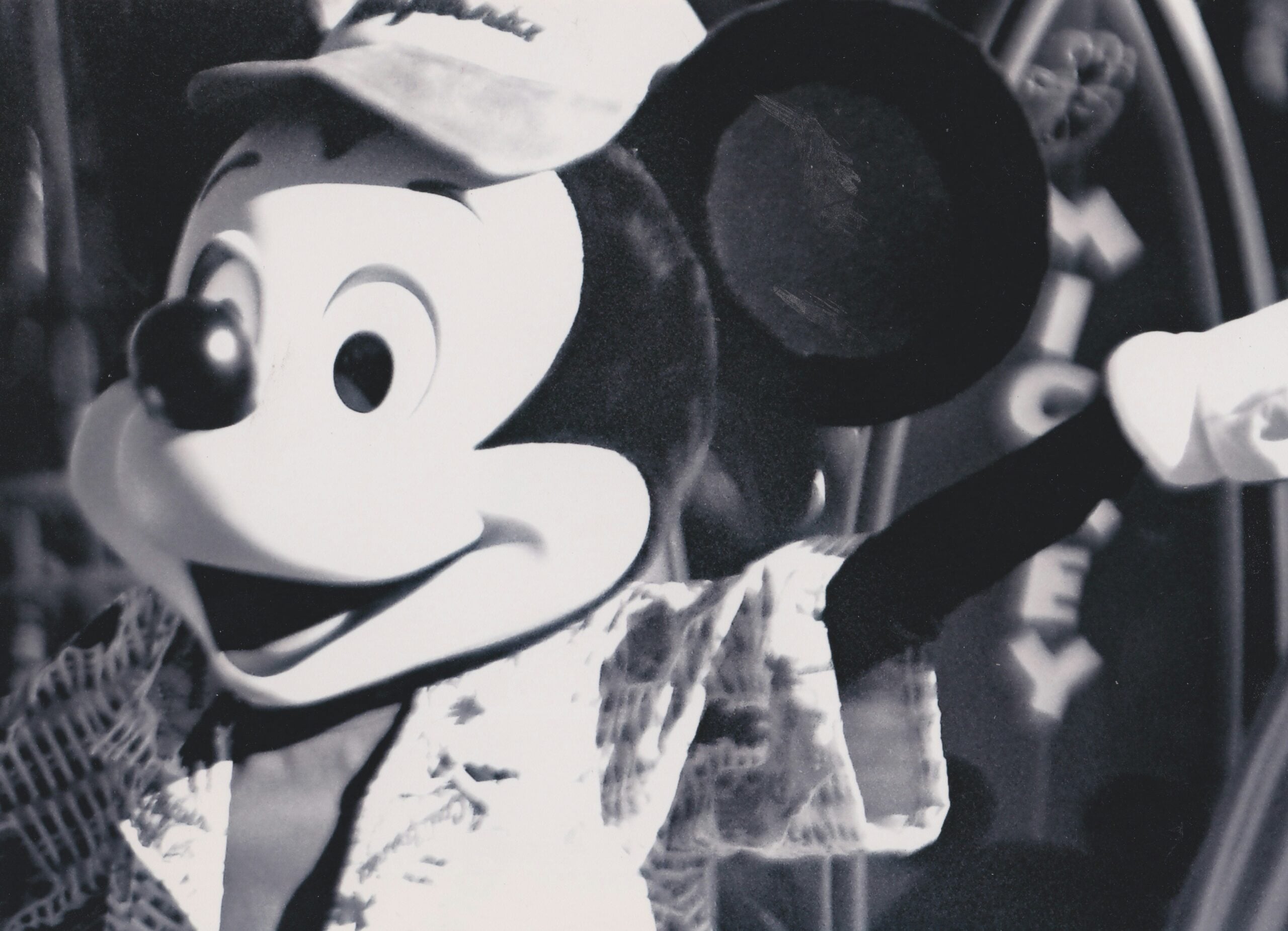 Black and white photo of a person in a Mickey Mouse costume wearing a hat and patterned shirt, smiling and waving, with part of a sign visible in the background.