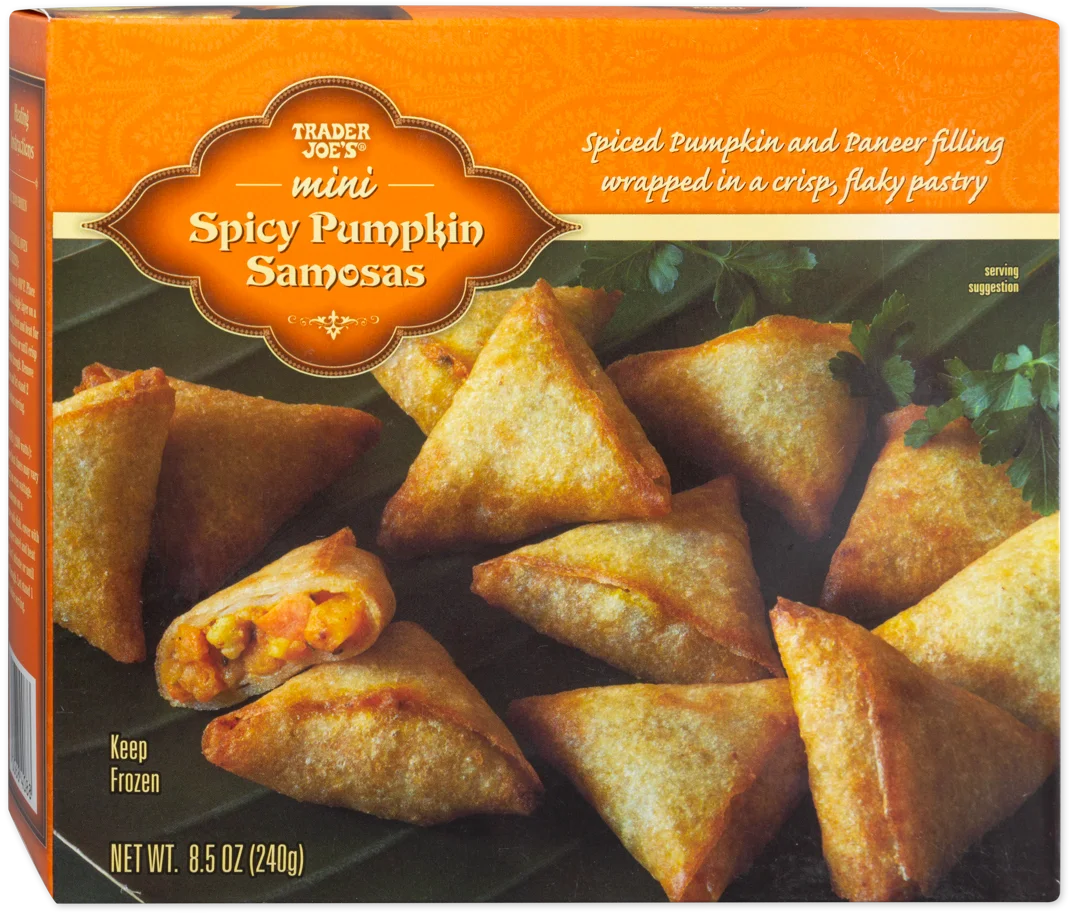Box of Trader Joe’s Mini Spicy Pumpkin Samosas, showing golden-brown, triangular pastries with visible spiced filling, arranged on a green surface with some cilantro garnish.