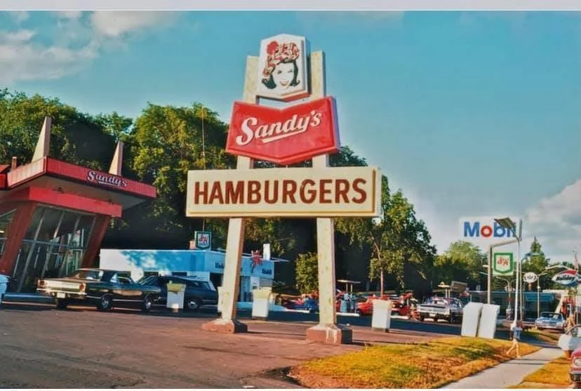 A vintage Sandy’s Hamburgers sign stands in front of a retro fast-food drive-in with classic cars parked nearby, surrounded by trees and additional gas station signs.