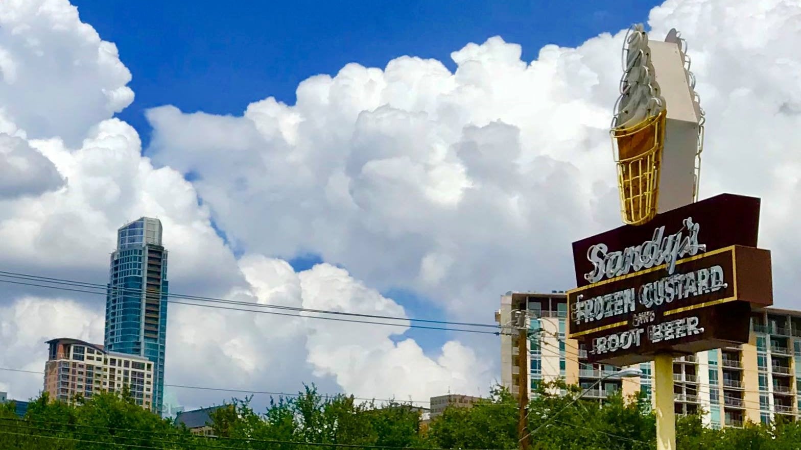 A retro sign for Sandy’s Frozen Custard and Root Beer stands against a backdrop of city buildings, a blue sky, and large fluffy clouds.