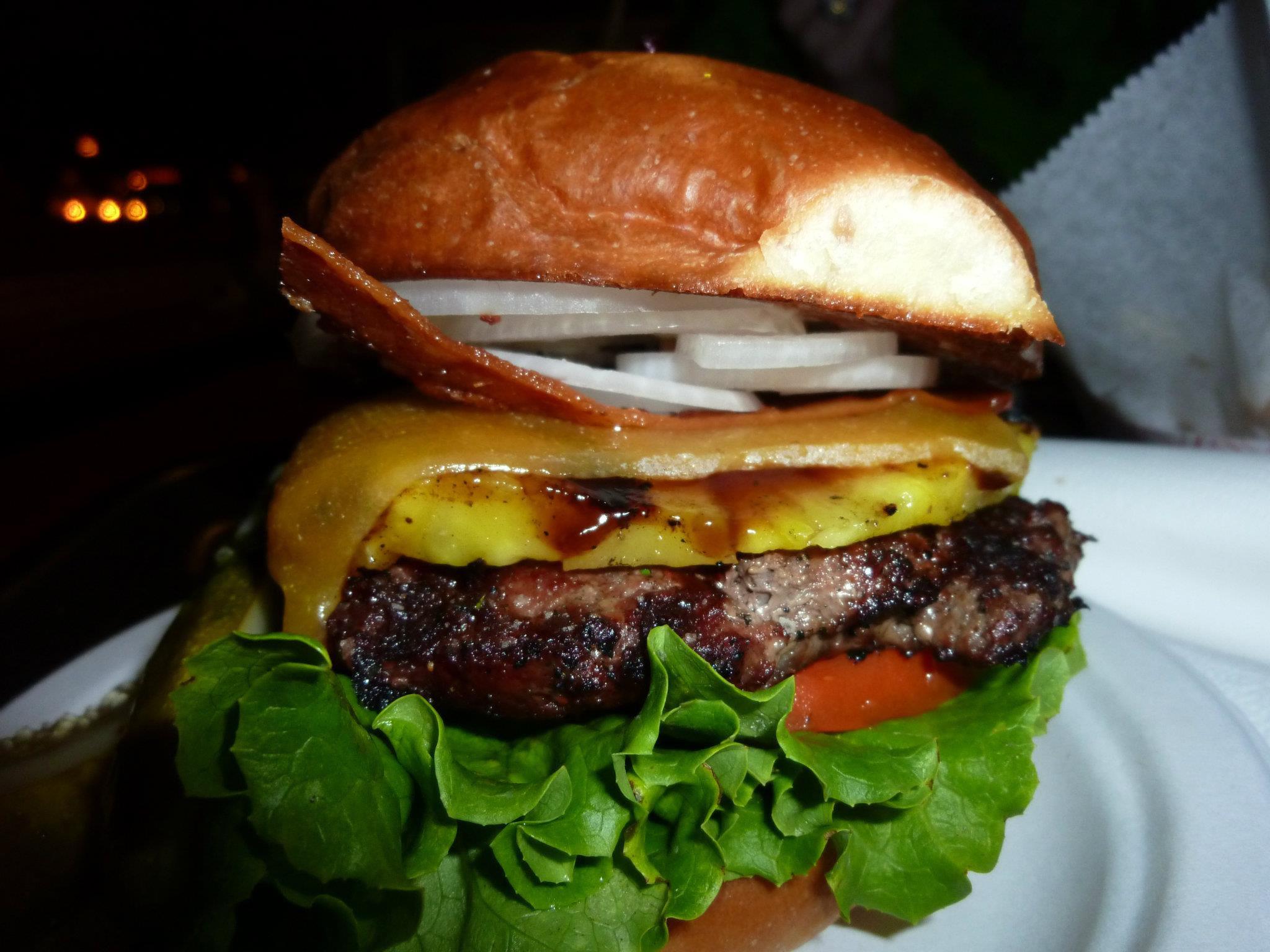 A close-up of a hamburger with lettuce, tomato, grilled pineapple, cheddar cheese, onions, bacon, and a glossy bun, served on a plate.