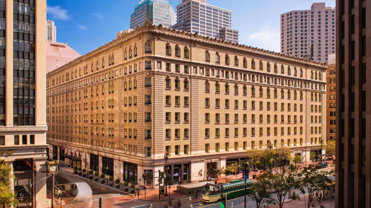 A large, historic beige building with many windows stands on a city corner, surrounded by tall modern skyscrapers and tree-lined streets. Reminiscent of the grand Historic Hotels found in bustling urban centers, the building offers a glimpse into the city’s storied past. A green trolley is visible on the street below.