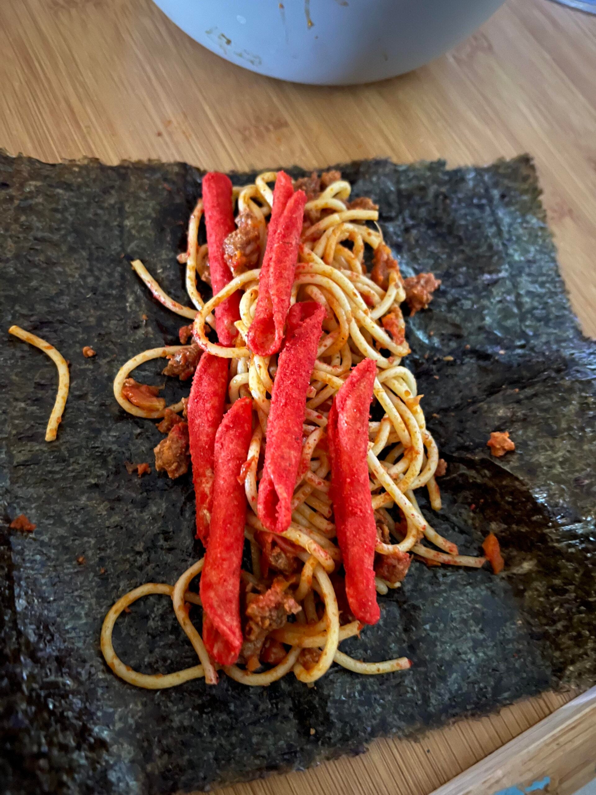 A sheet of seaweed topped with spaghetti and several red spicy snack sticks on a wooden surface.