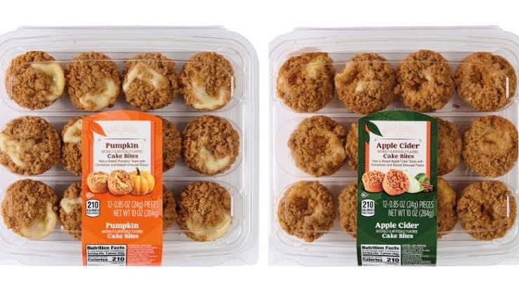 Two clear plastic packs of cake bites: one labeled “Pumpkin Cake Bites” with orange accents, the other “Apple Cider Cake Bites” with green accents. Both packs show twelve bites with crumb toppings. Fall grocery treats. 
