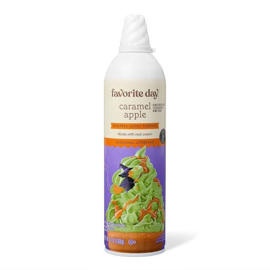 A can of Favorite Day caramel apple dairy topping with a white cap. The label shows green whipped topping, caramel drizzle, and a small witch illustration, indicating a seasonal offering.