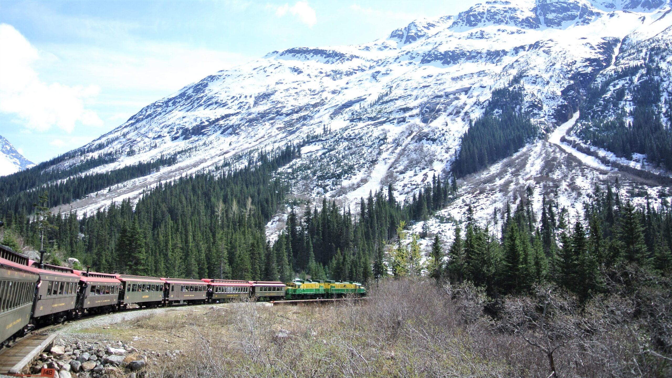 A vintage train travels through a forested valley with snow-covered mountains in the background under a partly cloudy sky.