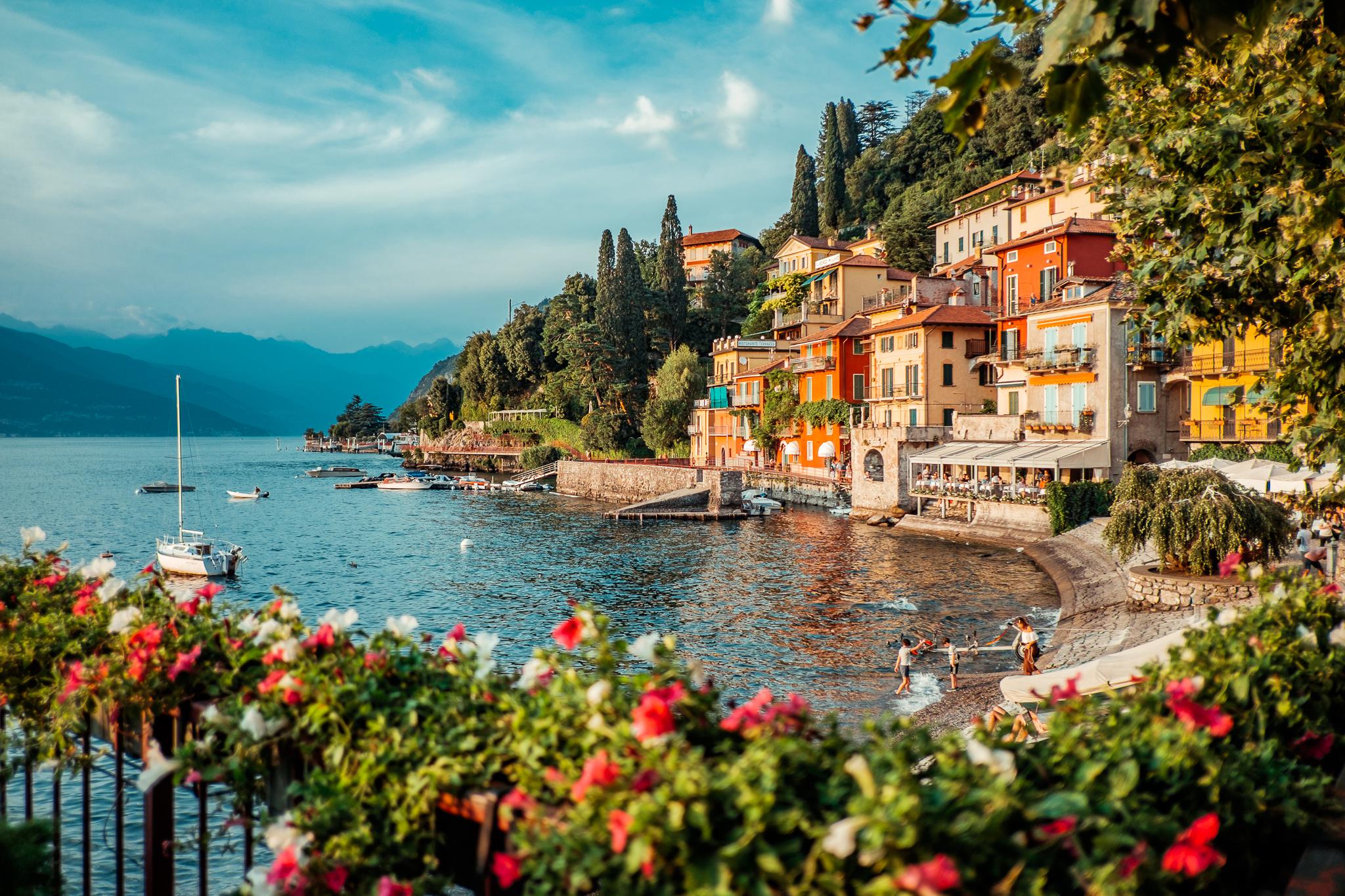 Colorful houses line the shore of a peaceful lake with boats, flowers in the foreground, and people enjoying the water under a bright, blue sky with hills in the background.