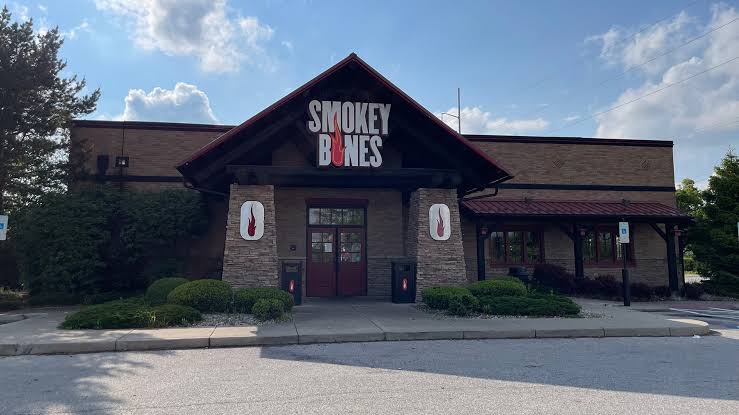 The exterior of a Smokey Bones restaurant with a large sign above the entrance, stone pillars, and neatly landscaped bushes in front under a partly cloudy sky. Restaurant chains on delivery apps.