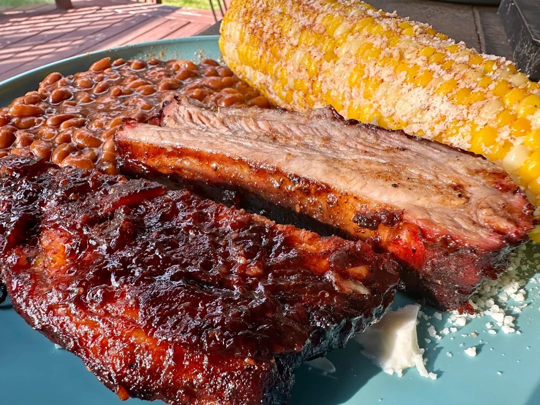 A plate of baked beans, barbecued ribs, and a buttered ear of corn topped with grated cheese, served on a blue plate.