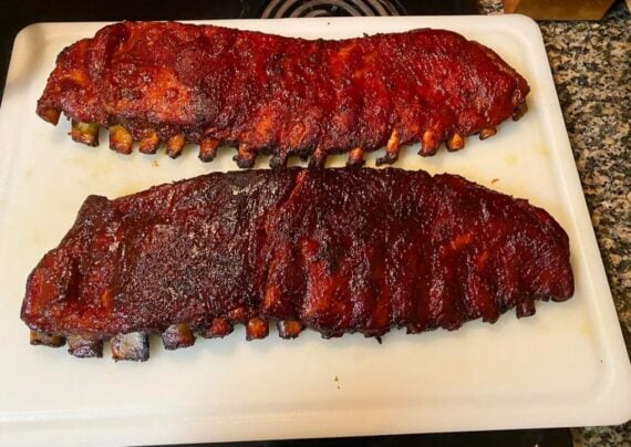 Two racks of barbecued ribs, coated in a dark, glossy sauce, sit side by side on a white cutting board on a kitchen counter.