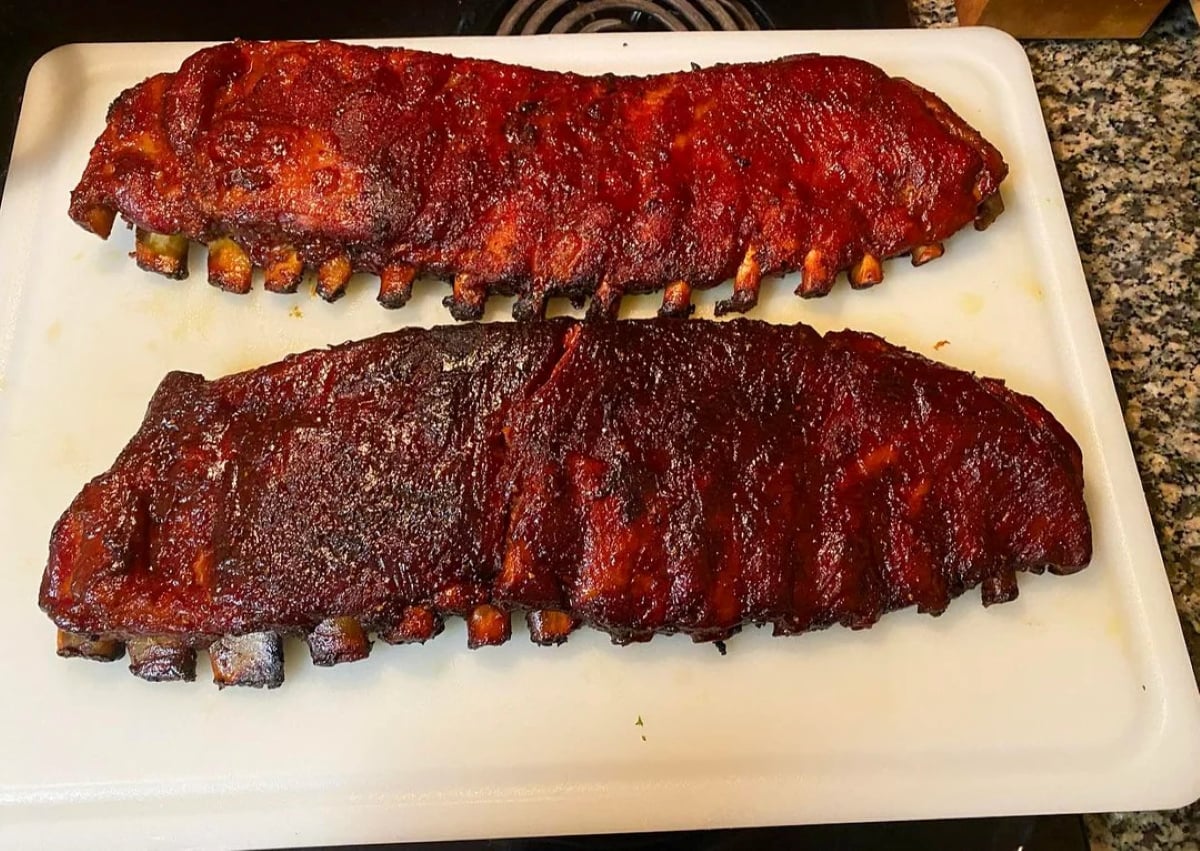 Two racks of barbecued ribs, coated in a dark, glossy sauce, sit side by side on a white cutting board on a kitchen counter.
