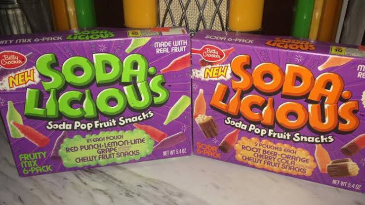 Two boxes of Betty Crocker Soda-Licious Soda Pop Fruit Snacks, one purple (fruity mix) and one orange (soda flavors), are placed side by side on a counter. Each box features images of colorful gummy snacks.