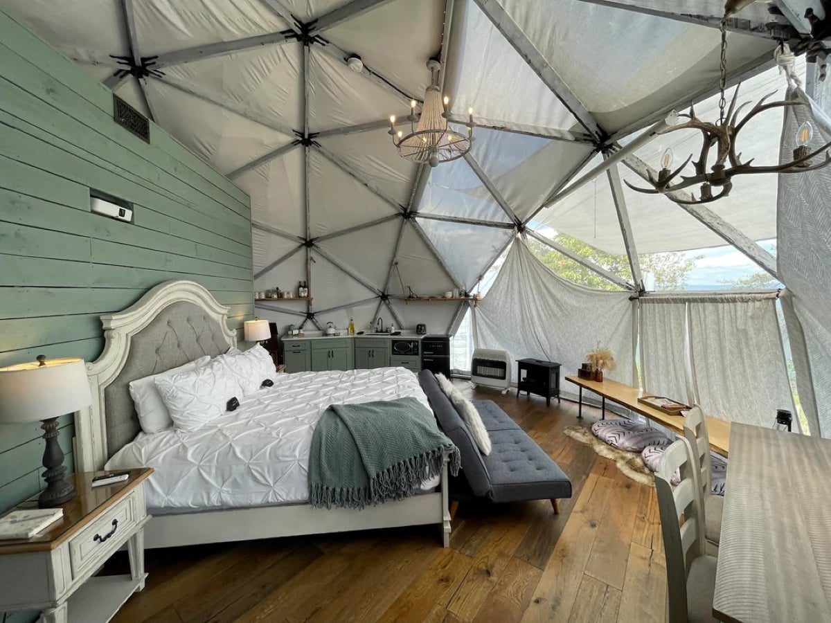 A cozy dome-shaped bedroom with a plush bed, green paneled wall, wood floors, a kitchenette, and large windows letting in natural light. Decor includes antler and chandelier lights.