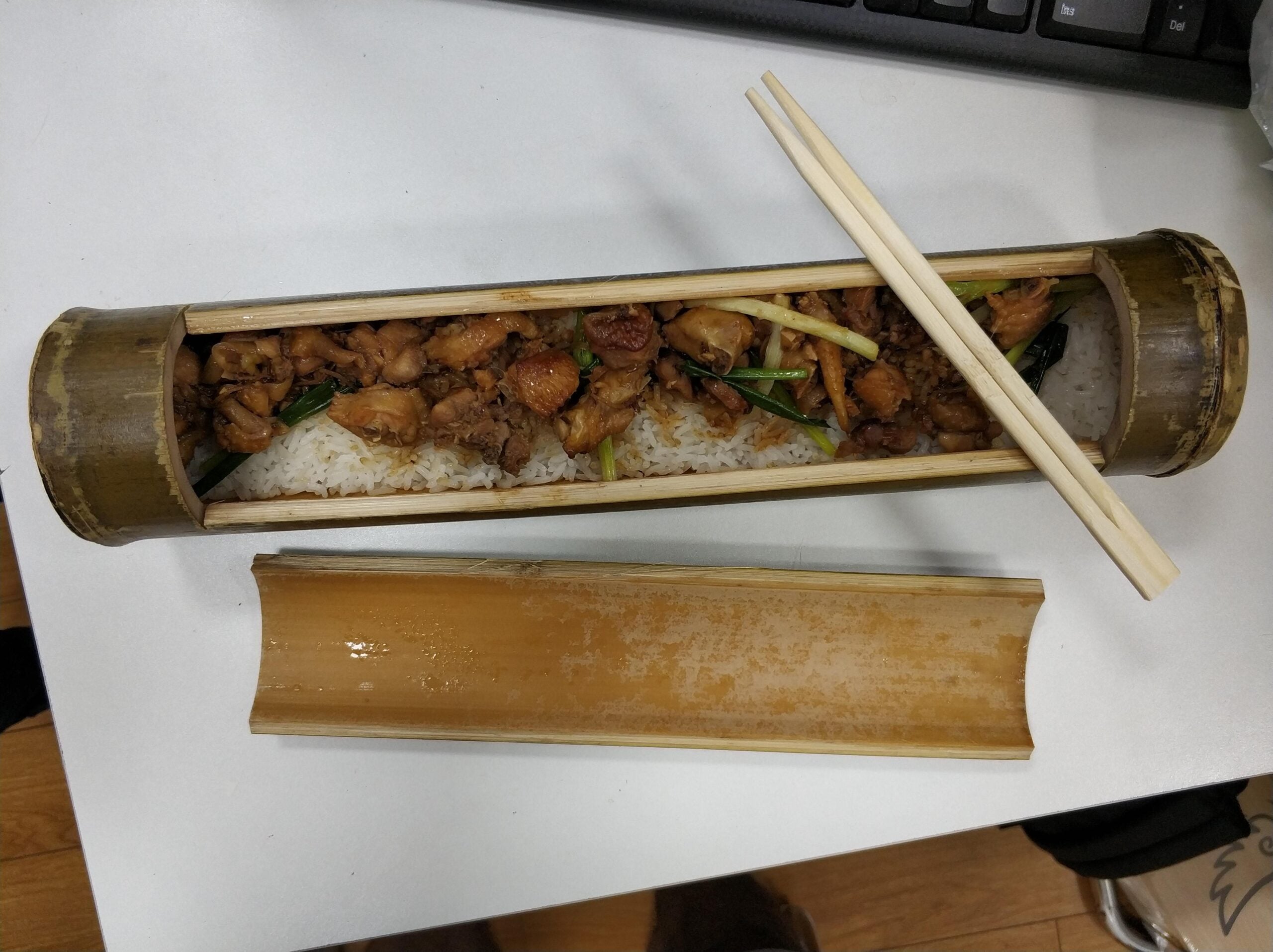 A bamboo tube filled with rice and stir-fried meat, served open on a white table with chopsticks resting on top.