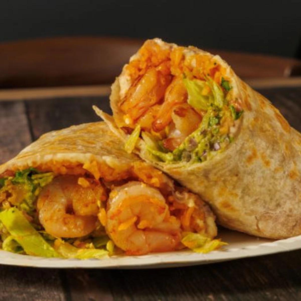 A plate with two halves of a shrimp burrito, filled with shrimp, lettuce, shredded cheese, and guacamole, on a wooden table.