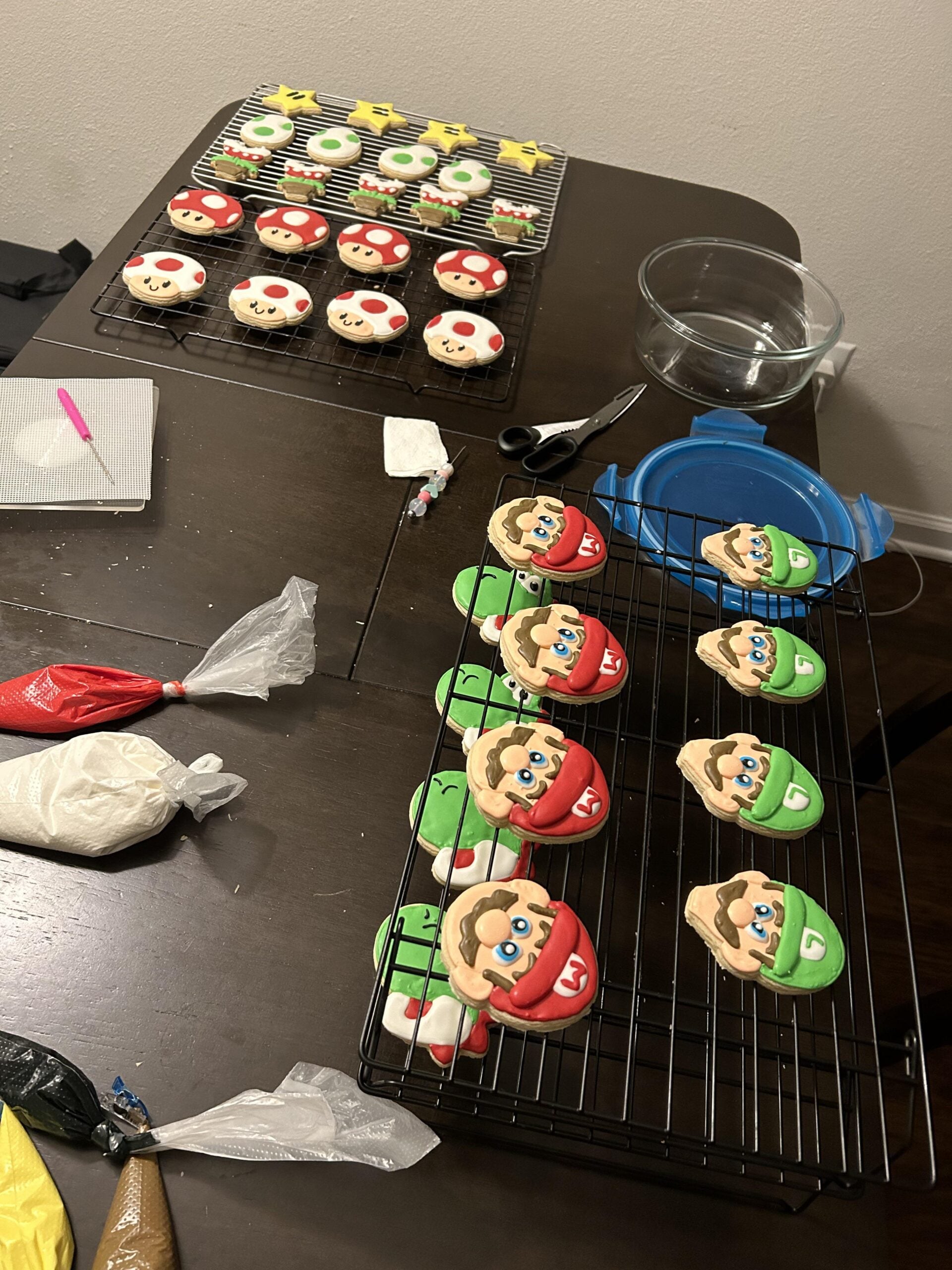 Cookies decorated as Mario, Luigi, Toad, and Super Stars from the Mario series are on cooling racks on a table with decorating supplies, piping bags, and containers nearby.