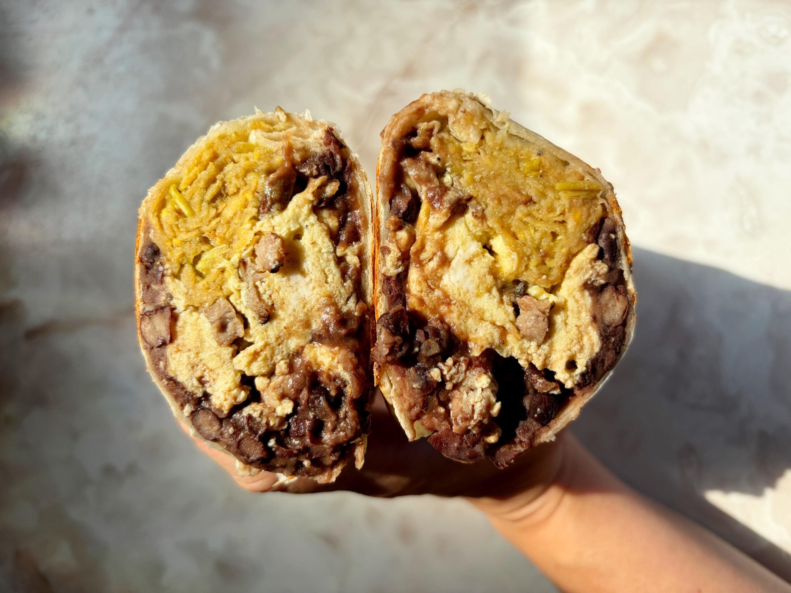 A hand holds a breakfast burrito cut in half, revealing layers of scrambled eggs, black beans, shredded potatoes, and possibly sausage inside a flour tortilla.