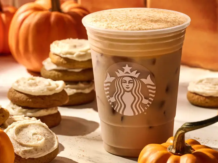 A Starbucks iced coffee with a dusting of spices sits among frosted cookies and small pumpkins, evoking an autumn theme.
