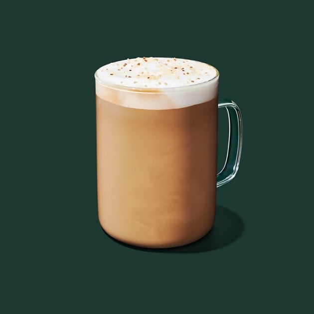 A clear glass mug filled with a frothy, light brown latte topped with foam and a sprinkle of spices, set against a solid dark green background.