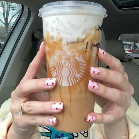 Person holding a large Starbucks iced banana bread latte in a car; nails are painted pink with red hearts, and the drink has whipped cream on top.