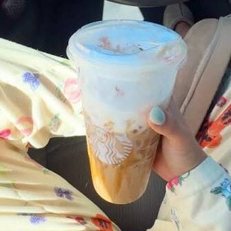 A person in floral pajamas sits in a car, holding a large iced coffee with a plastic lid and straw. Their nails are painted light blue, and the Starbucks logo is visible on the cup. 15 Starbucks Fall Menu Items to Try in 2026.