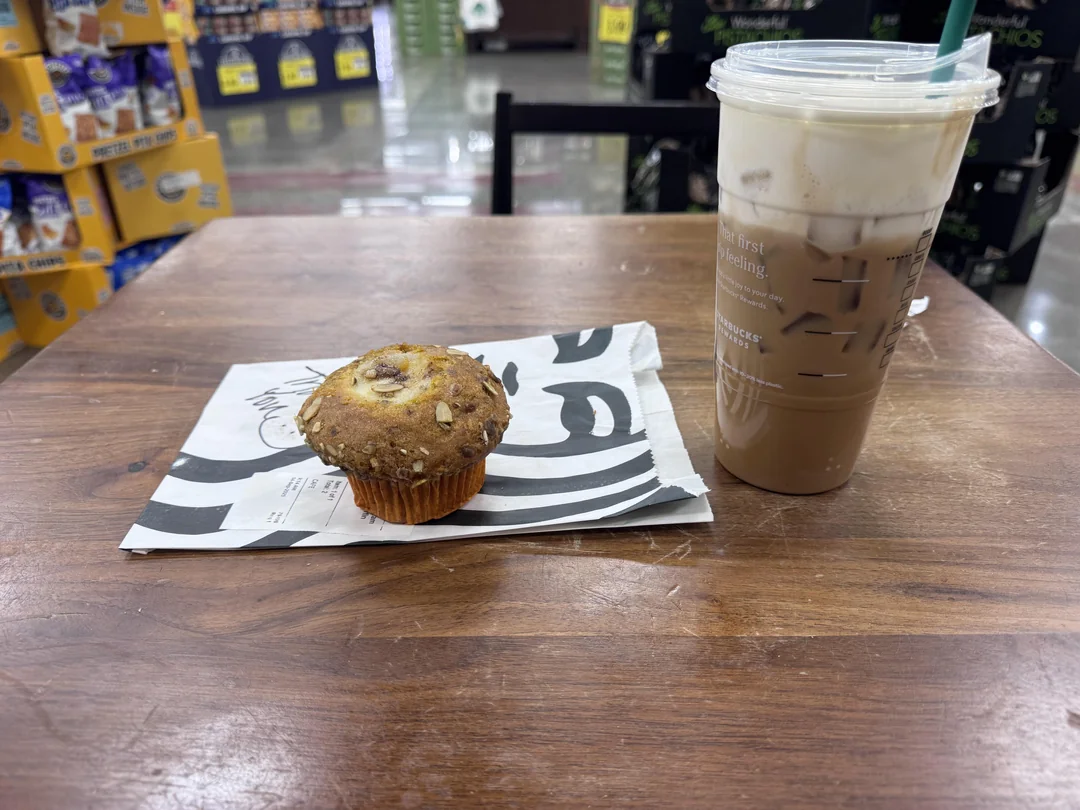 A chocolate chip muffin on a napkin and a plastic cup of iced coffee with a green straw sit on a wooden table in a grocery store cafe area.