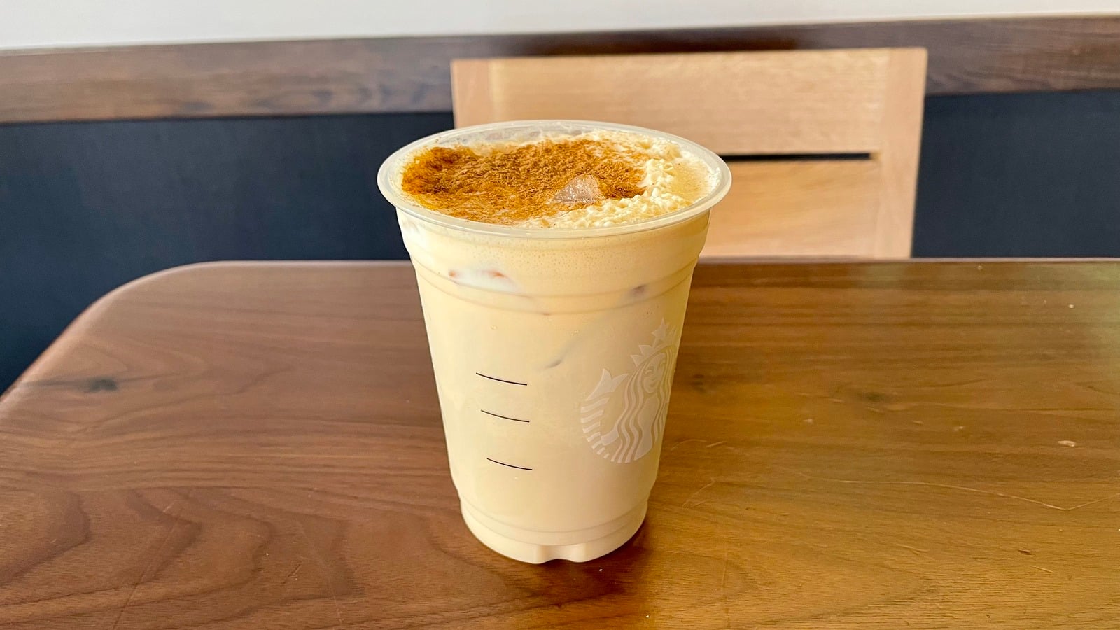 A plastic cup of iced coffee topped with foam and sprinkled cinnamon sits on a wooden table in front of a wooden chair.