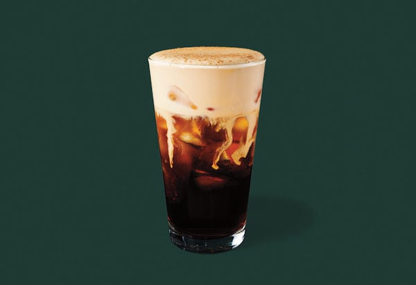 A glass of iced coffee topped with creamy foam sits against a dark green background. The coffee is dark with visible ice cubes and a layer of light, frothy cream on top.