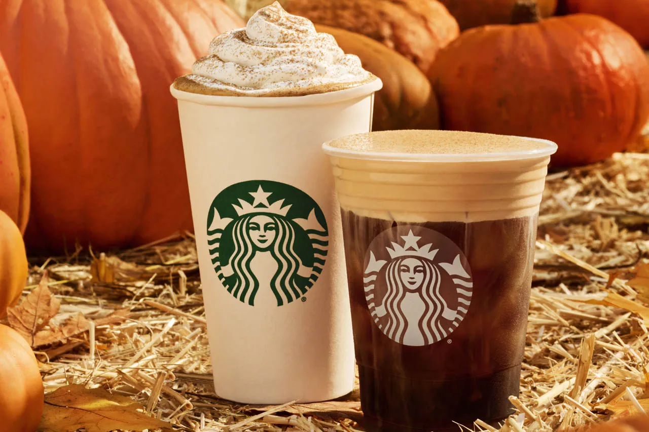 Two Starbucks drinks, one hot with whipped cream and one iced, are set on straw among pumpkins, evoking an autumn theme.