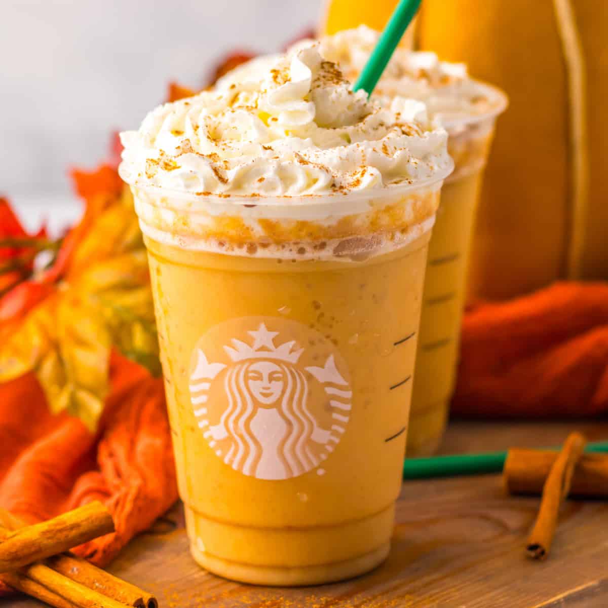 A Starbucks pumpkin spice frappuccino topped with whipped cream and sprinkled spices, with a green straw, sits on a wooden surface surrounded by cinnamon sticks and autumn leaves.