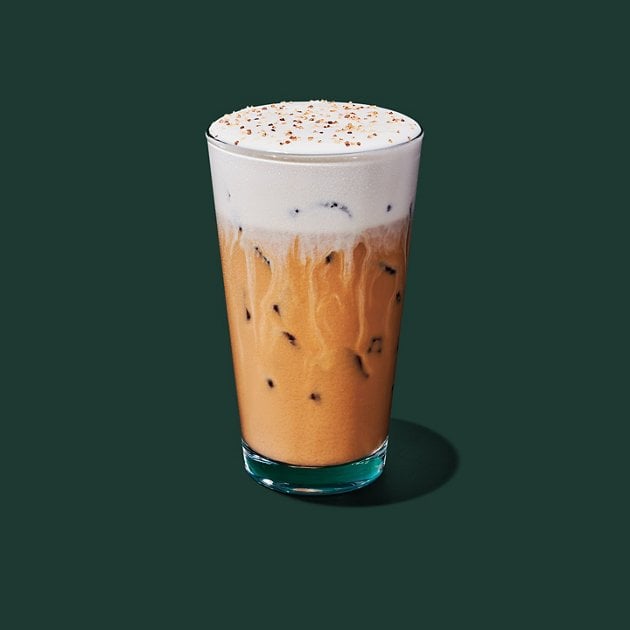 A glass of iced coffee with a layer of creamy foam and sprinkles on top, against a solid dark green background.