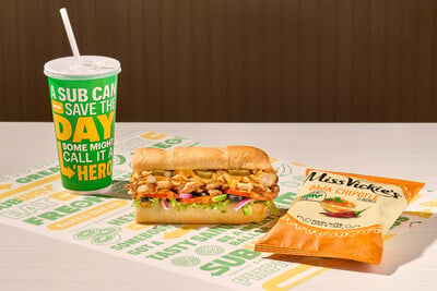 A fountain drink, a sub sandwich with lettuce, pickles, and chicken, and a bag of Miss Vickie’s Baja Chipotle chips sit on a table with Subway-branded paper and background.