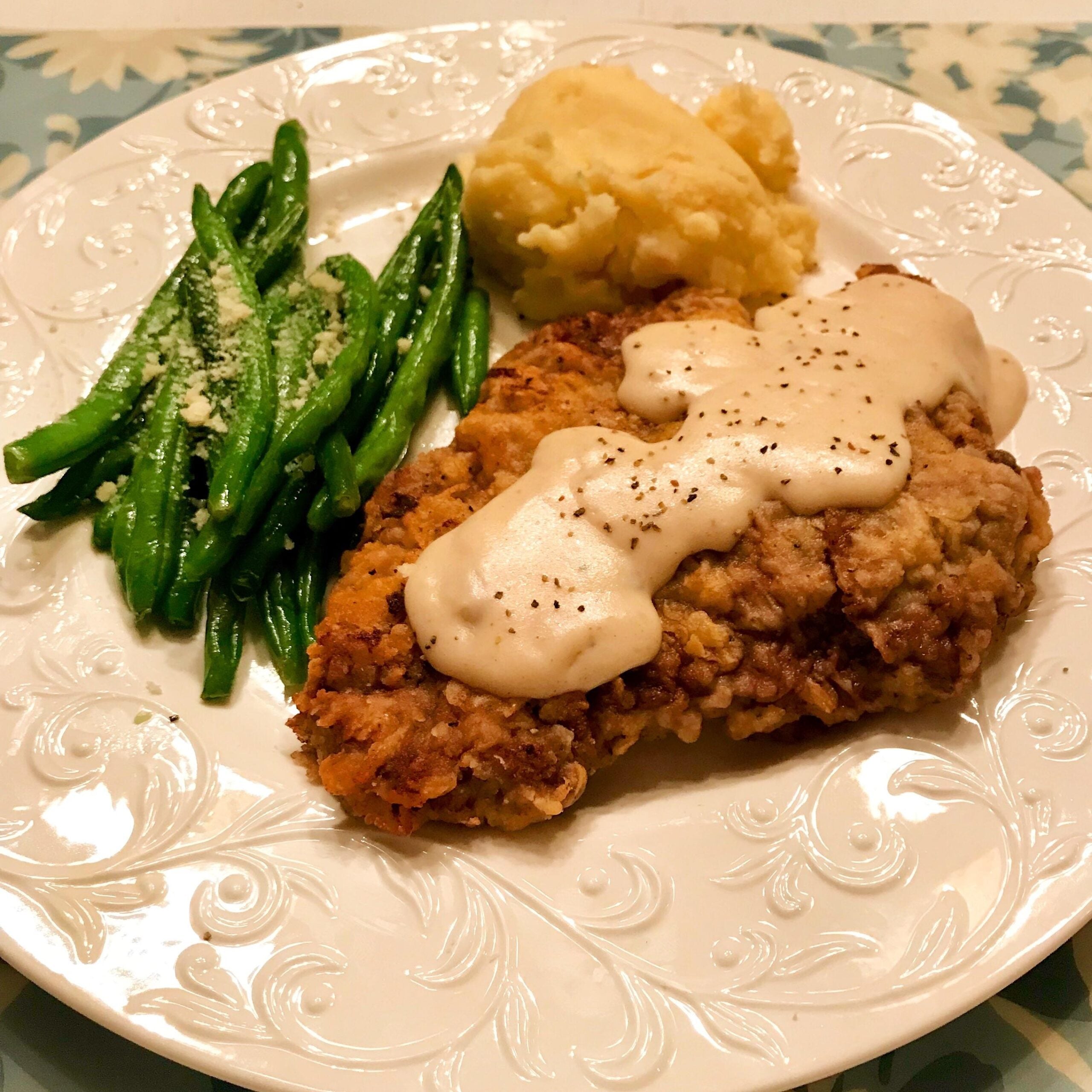 A plate with fried chicken topped with white gravy, a serving of mashed potatoes, and green beans sprinkled with grated cheese.