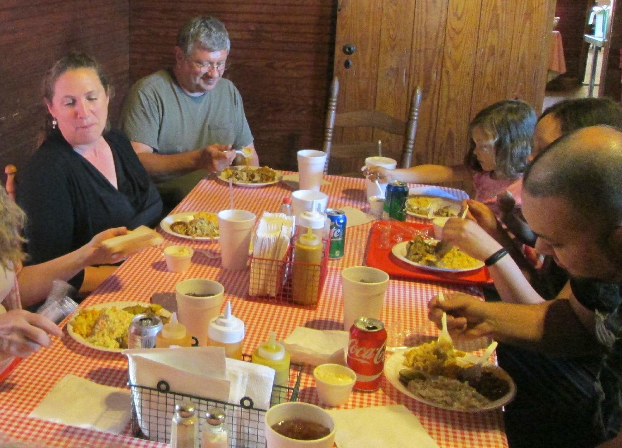 Six people sit around a red-checked table eating a meal. Various plates of food and drinks, including a can of Coke, are visible. The group appears to be enjoying lunch together in a cozy setting.