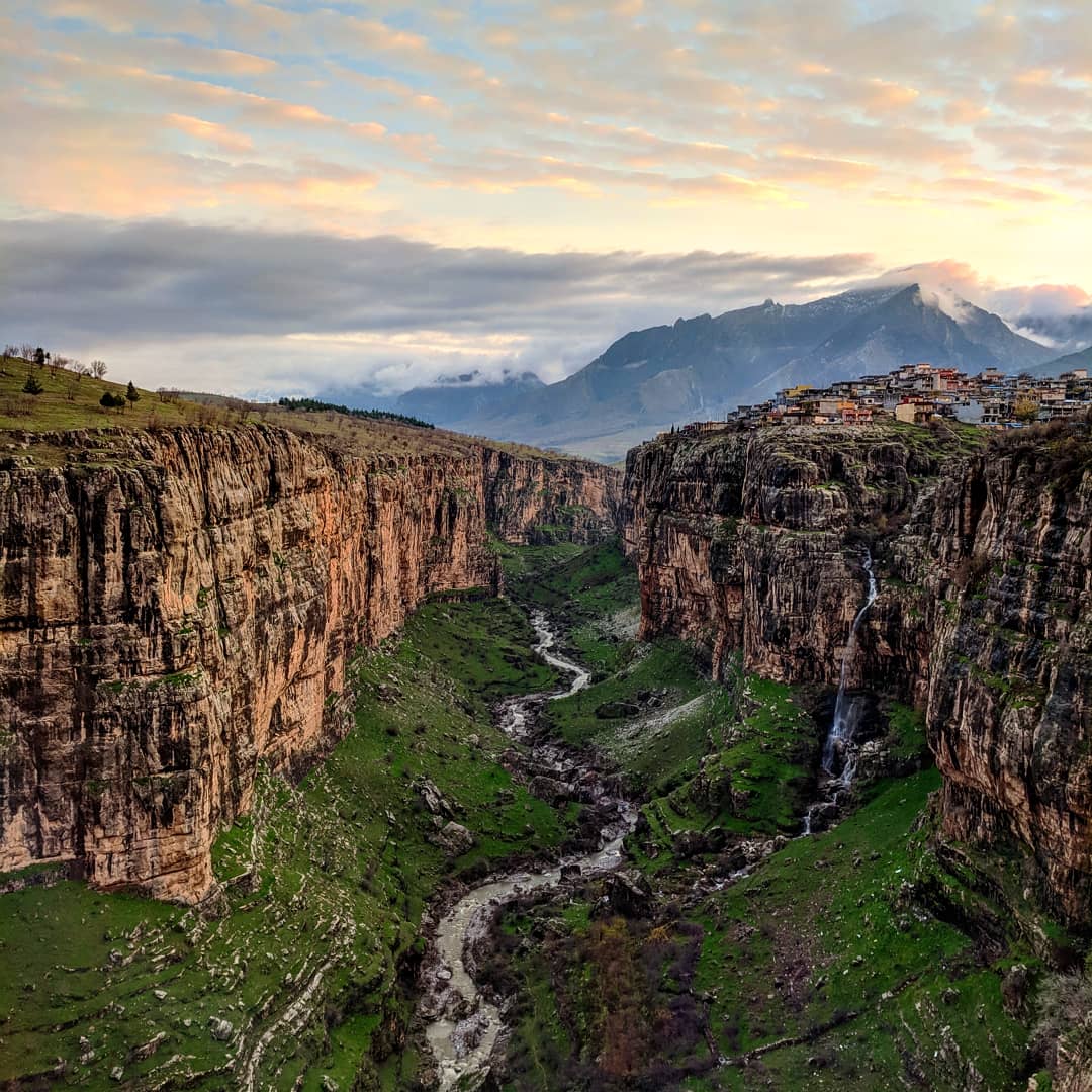 A deep green valley with a river runs between steep, rocky cliffs; a small village sits atop the right cliff. Mountains and clouds appear in the background at sunset.