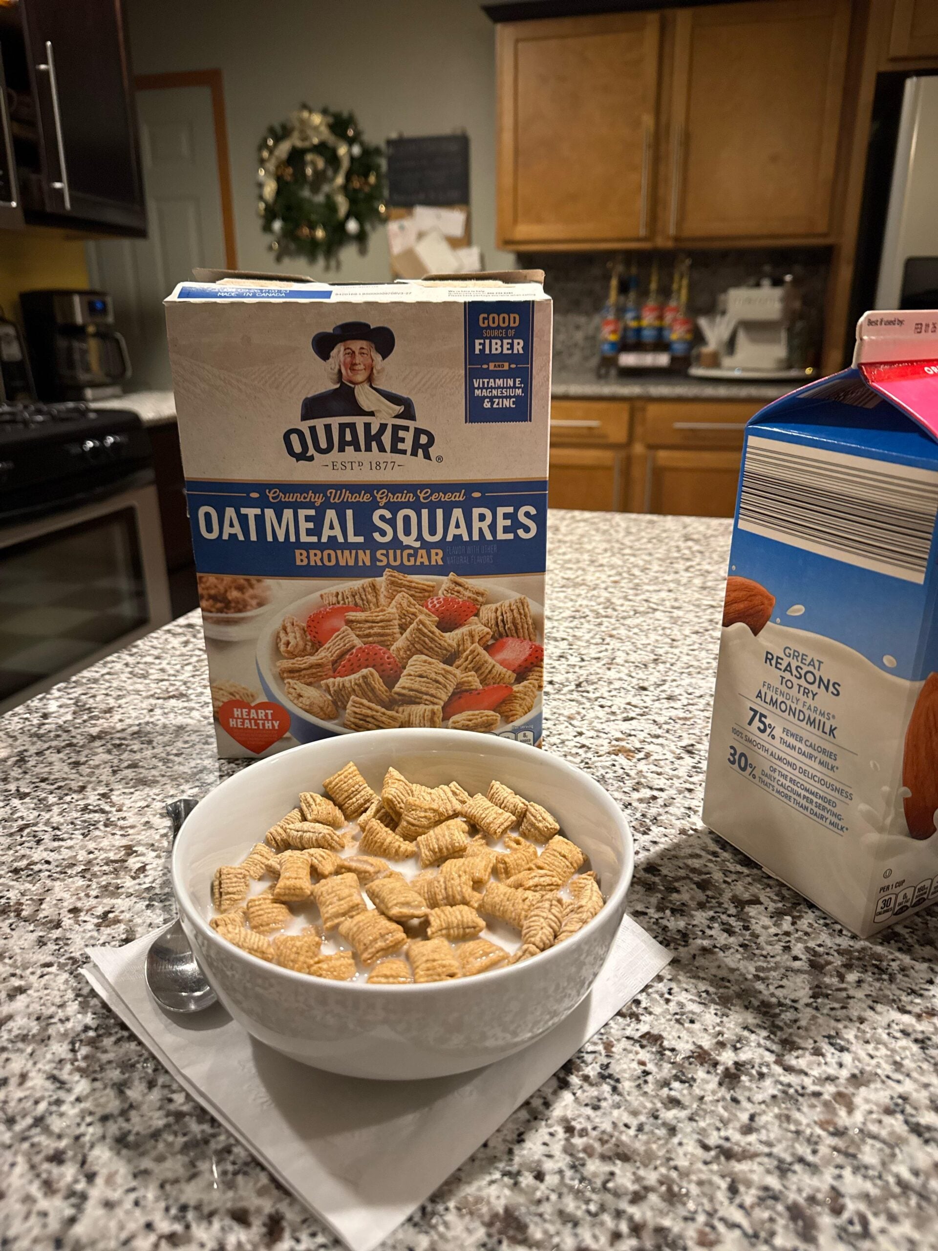 A bowl of Quaker Oatmeal Squares cereal with milk sits on a kitchen counter. The cereal box and a carton of almond milk are nearby, with wooden cabinets and kitchen appliances in the background.