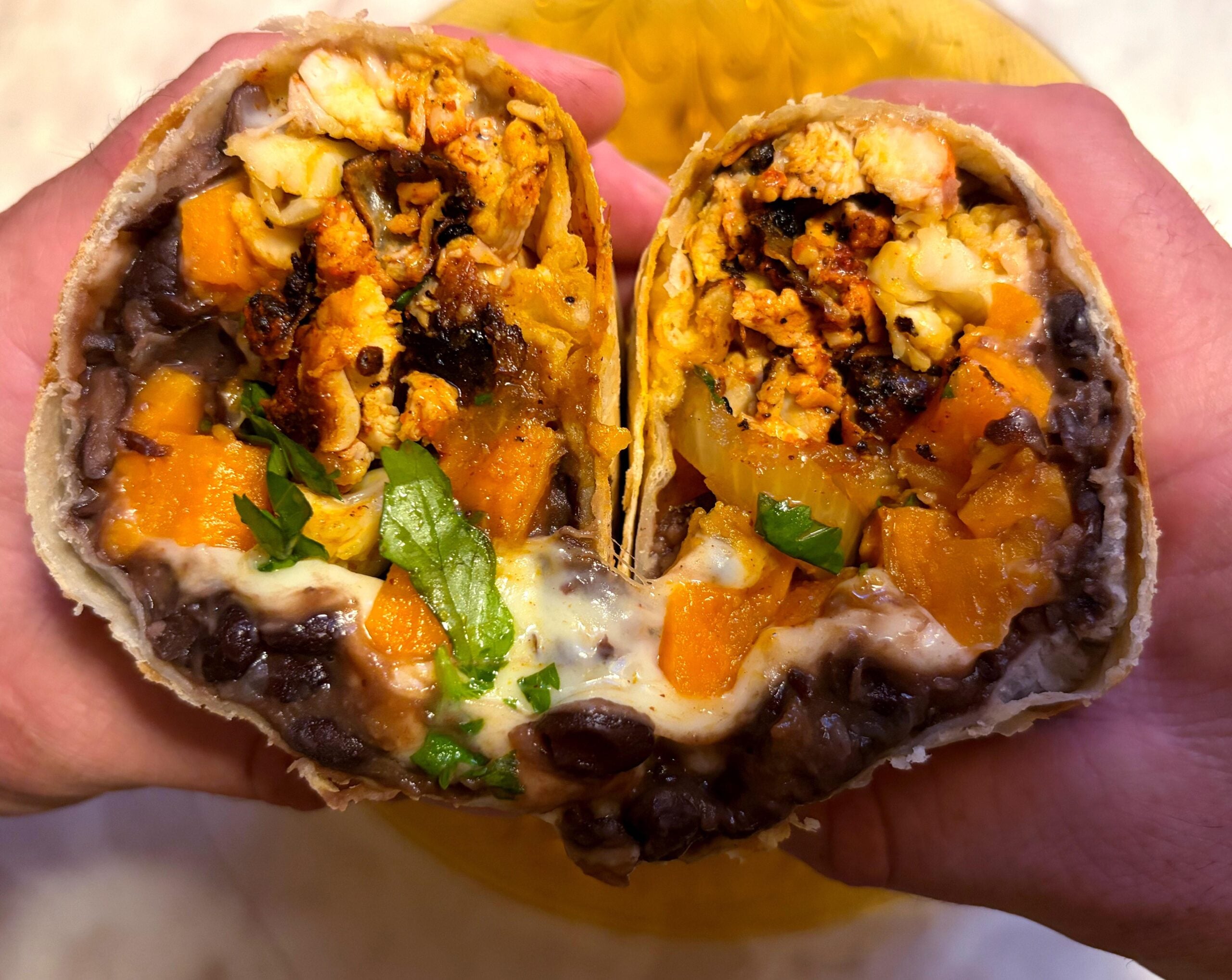 Two hands hold a burrito cut in half, revealing black beans, melted cheese, roasted sweet potatoes, cauliflower, and fresh herbs wrapped in a tortilla.