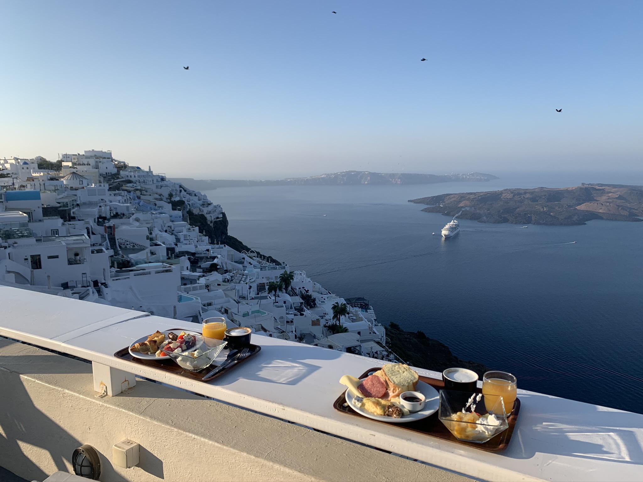 Breakfast trays with juice, fruit, and pastries on a white ledge overlook white buildings on a cliff, with the sea, a cruise ship, and blue sky in the background.